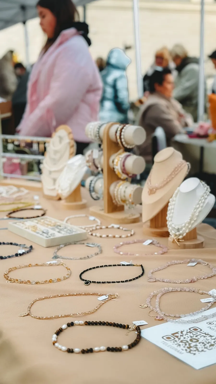 A jewelry pop-up table at a market, necklaces on stands with browsing customers softly out of focus